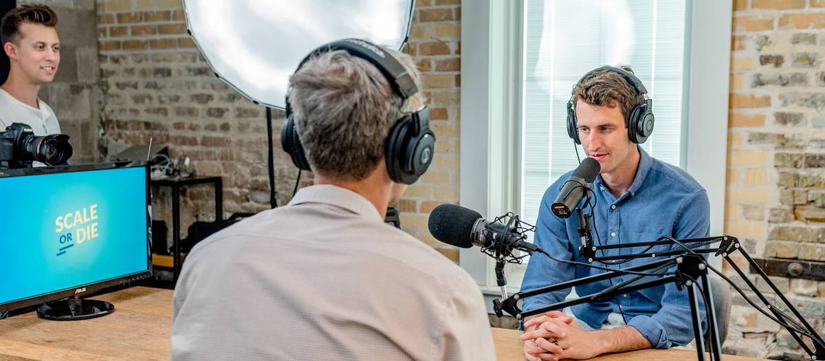 Two men being interviewed within a podcast studio. There is also a professional photographer on the left of the room.