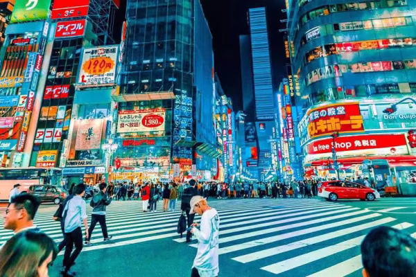 Busy city shopping strip at night with illuminated storefronts, neon signs, and shoppers walking along the sidewalk