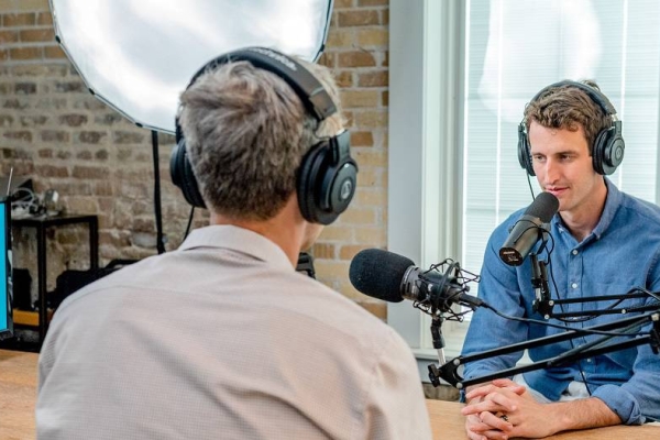 Two men being interviewed within a podcast studio. There is also a professional photographer on the left of the room.